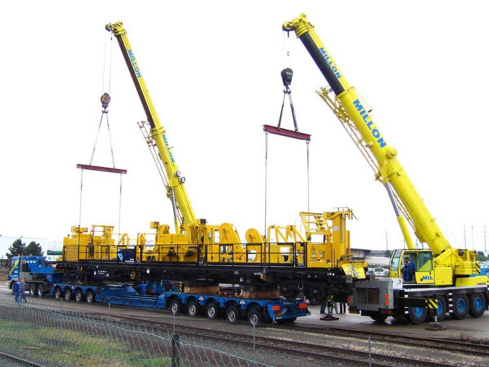 Deux grues mobiles jaunes travaillent de concert pour soulever une grande machine industrielle jaune d'une remorque à plateau à plusieurs essieux.