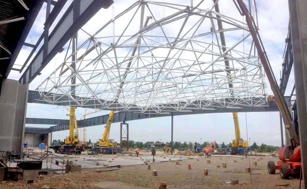Des grues soulèvent une grande structure de toit en acier blanc sur un chantier de construction.