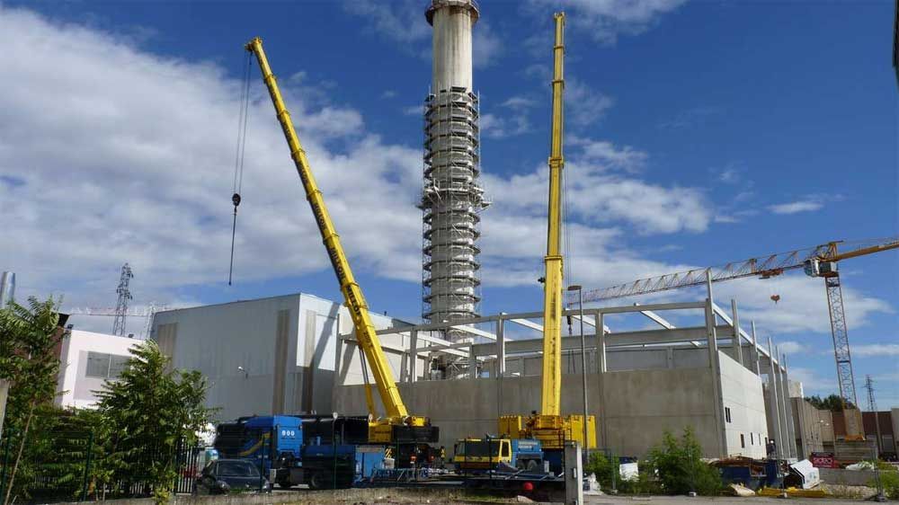 Deux grues jaunes sont stationnées devant un bâtiment industriel abritant une centrale électrique, surmonté d'une haute cheminée, sous un ciel bleu.