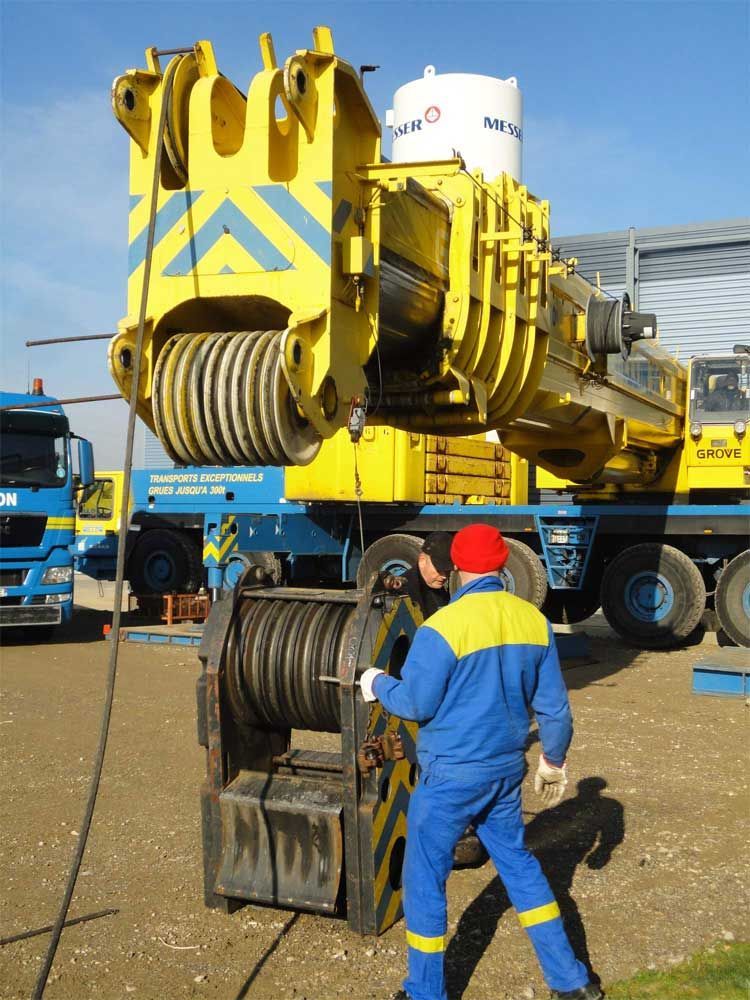 Un ouvrier en uniforme bleu et jaune aide à enfiler un crochet de grue robuste sur les câbles d'une grue mobile jaune.
