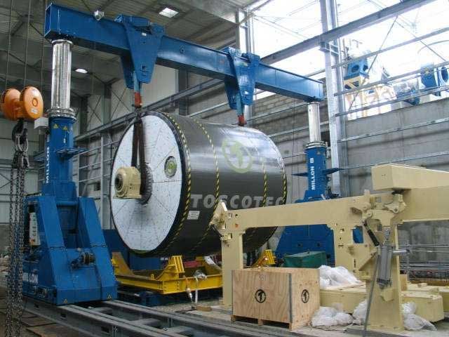 Un gros cylindre industriel marqué Toscotec est soulevé par un portique bleu à l'intérieur d'un entrepôt d'usine.