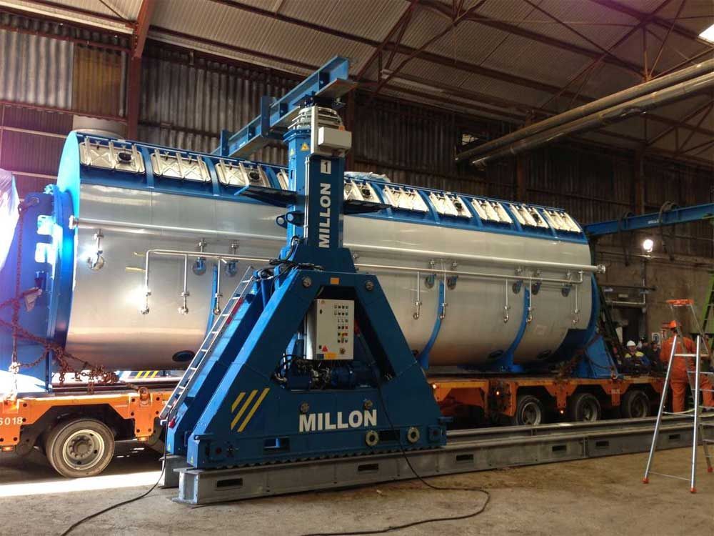 Une grande grue portique industrielle bleue Millon soulevant un réservoir cylindrique argenté dans un environnement d'usine.