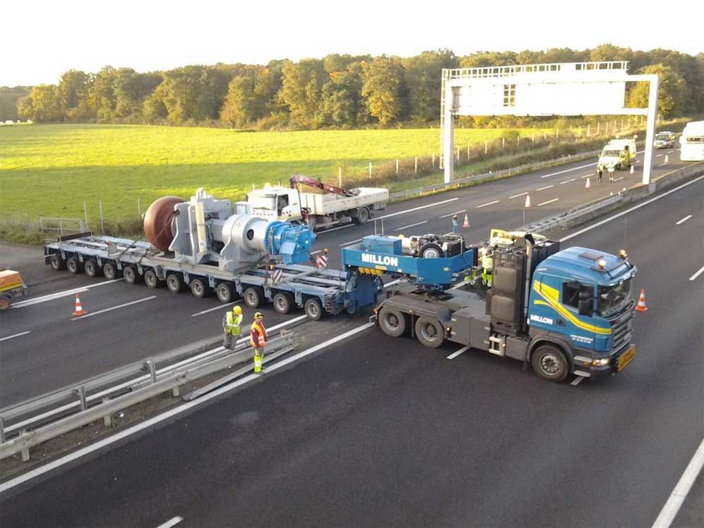 Un semi-remorque bleu transporte un composant industriel massif sur une remorque à plusieurs essieux, traversant une autoroute.