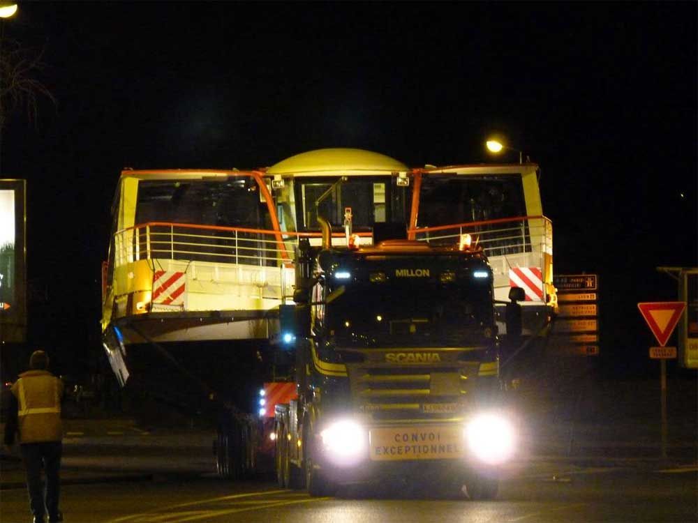 Une scène nocturne sombre montrant un camion poids lourd transportant une grande section de bateau jaune sur une large remorque surbaissée.
