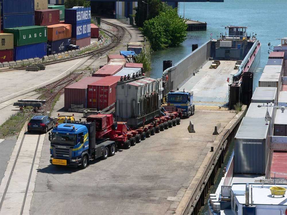 Un camion bleu transportant un gros transformateur industriel vers une barge dans un terminal maritime.