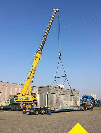 Sous un ciel bleu dégagé, une grue mobile jaune soulève une grande unité de climatisation industrielle d'une remorque à plateau.