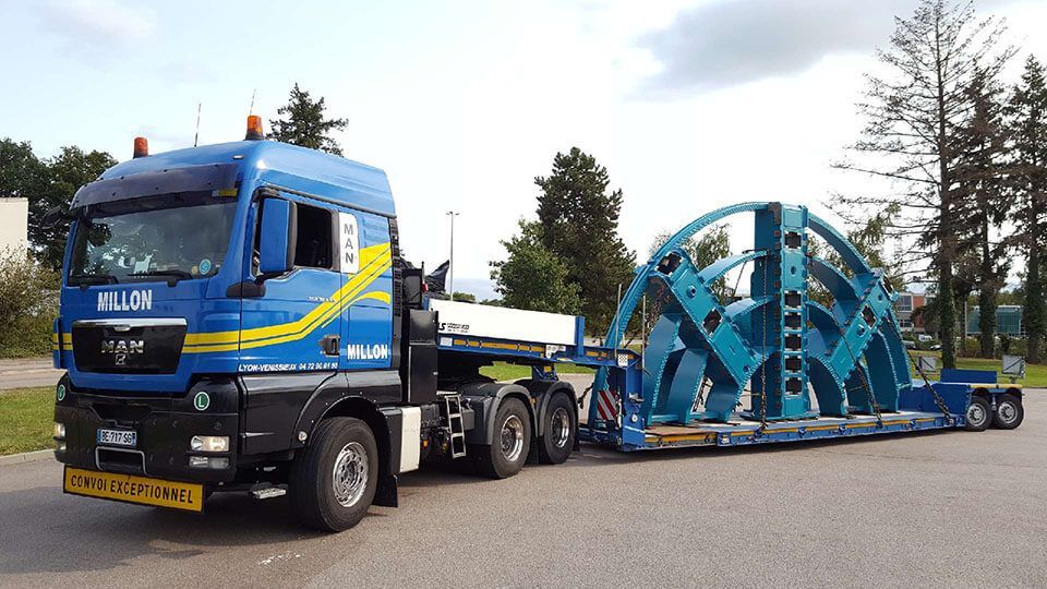 Un semi-remorque MAN bleu transportant une pièce de machine industrielle bleue surdimensionnée sur une remorque surbaissée, stationné sur un terrain goudronné.