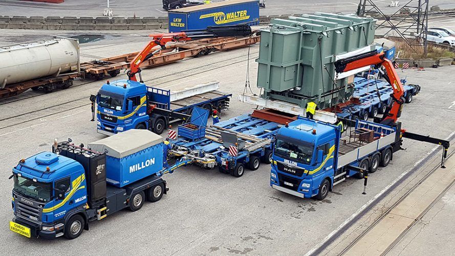 Deux camions bleus et une grue transportent un gros transformateur industriel vert sur une remorque robuste dans une cour de chargement.
