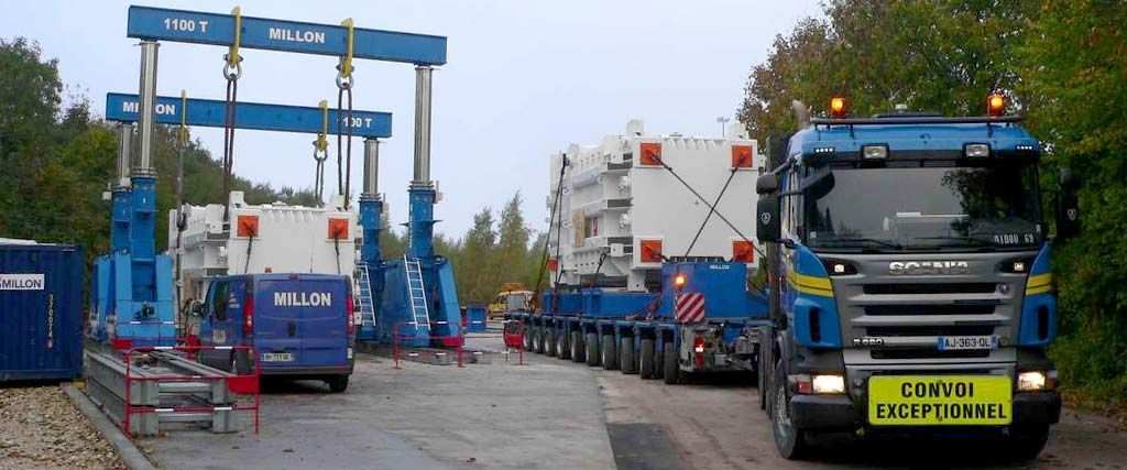 Un semi-remorque bleu transporte une importante cargaison industrielle près de deux gigantesques portiques bleus, dans une zone extérieure pavée.