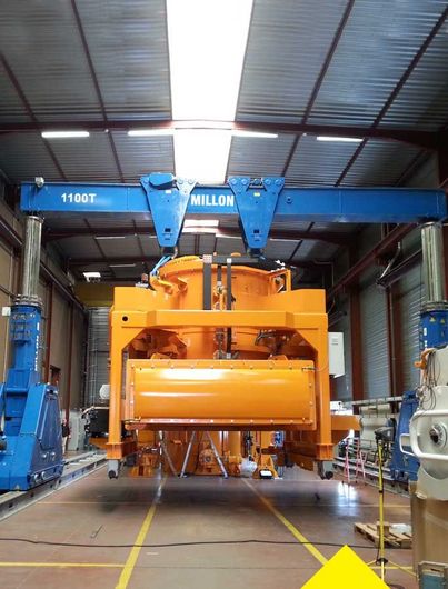 Une grue portique bleue robuste soulève une grande machine industrielle orange à l'intérieur d'un entrepôt spacieux.