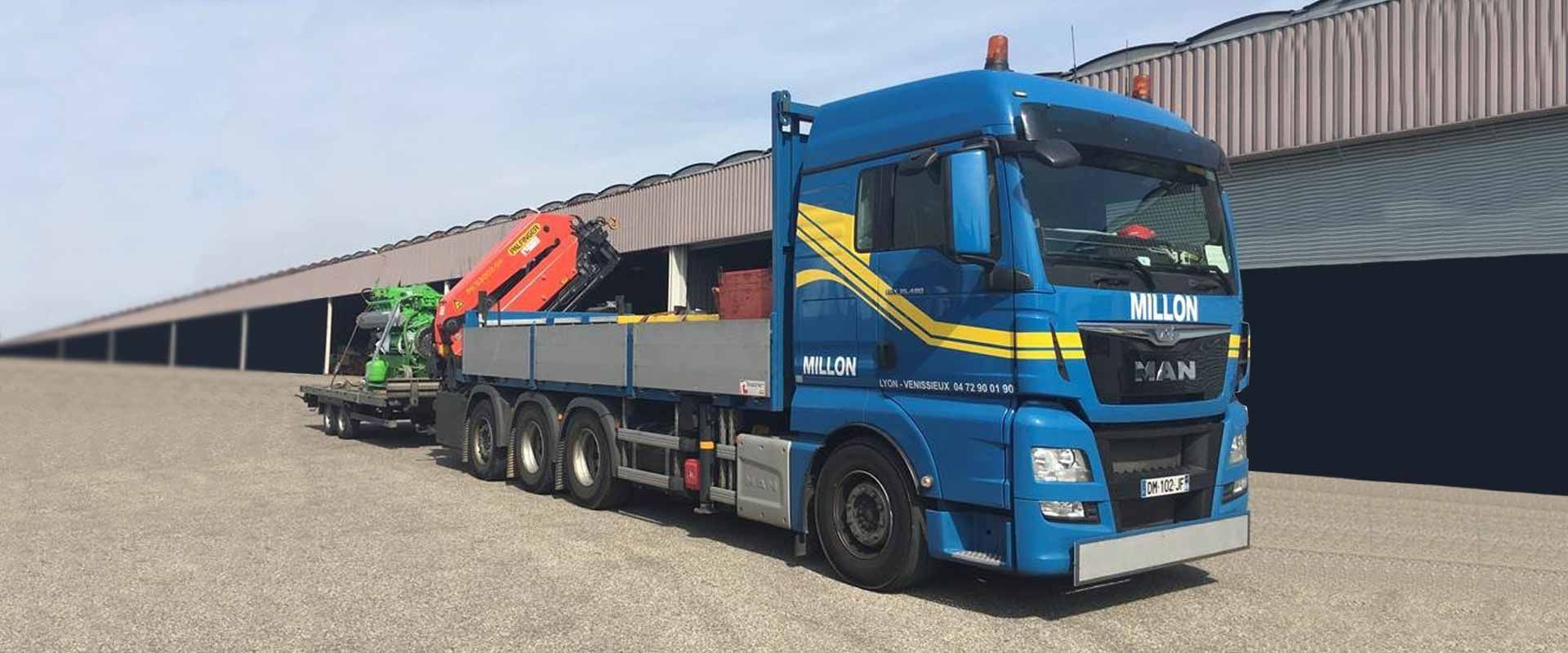 Un camion MAN bleu équipé d'une grue est stationné sur un terrain gravillonné devant un long entrepôt industriel.