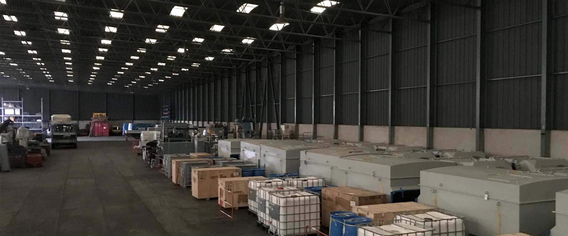 Un vaste entrepôt industriel présentant des rangées de marchandises emballées en caisses, en boîtes et sur palettes, sur un immense sol en béton sombre.