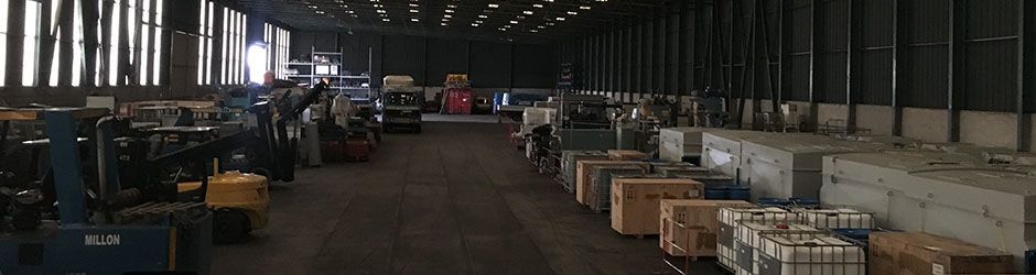 Vue grand angle d'un vaste entrepôt faiblement éclairé, avec des machines industrielles et diverses caisses en bois au sol.