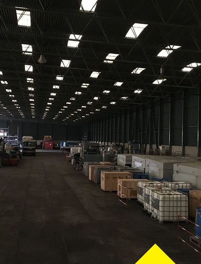 Un entrepôt industriel spacieux et faiblement éclairé, avec de hauts plafonds percés de puits de lumière et des rangées de caisses stockées à même le sol.