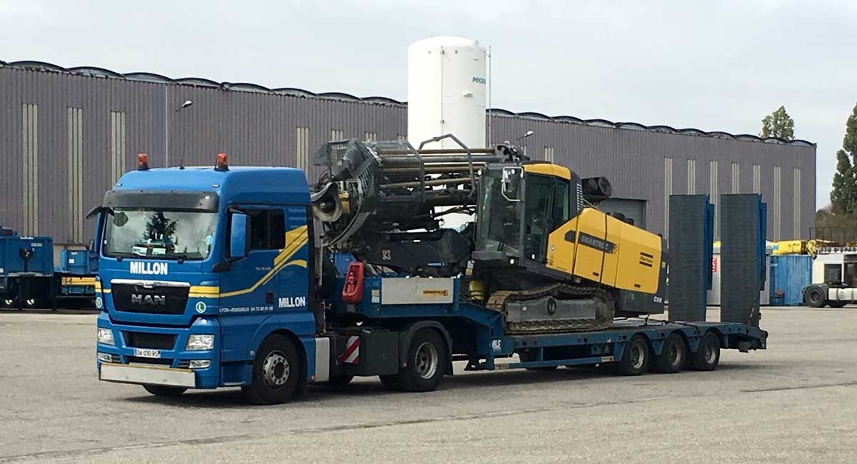 Un semi-remorque MAN bleu transportant une foreuse chenillée jaune sur une remorque surbaissée dans une cour industrielle pavée.