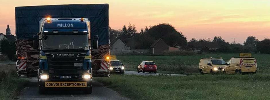 Un camion bleu hors gabarit circule sur une route au coucher du soleil, escorté par plusieurs voitures pilotes en arrière-plan.