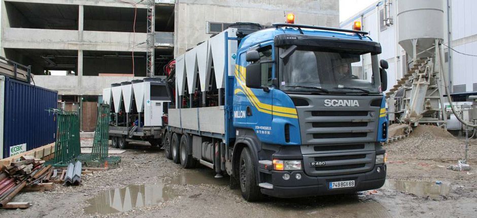 Un camion Scania bleu avec une remorque transportant du matériel industriel sur un chantier de construction boueux et inachevé.