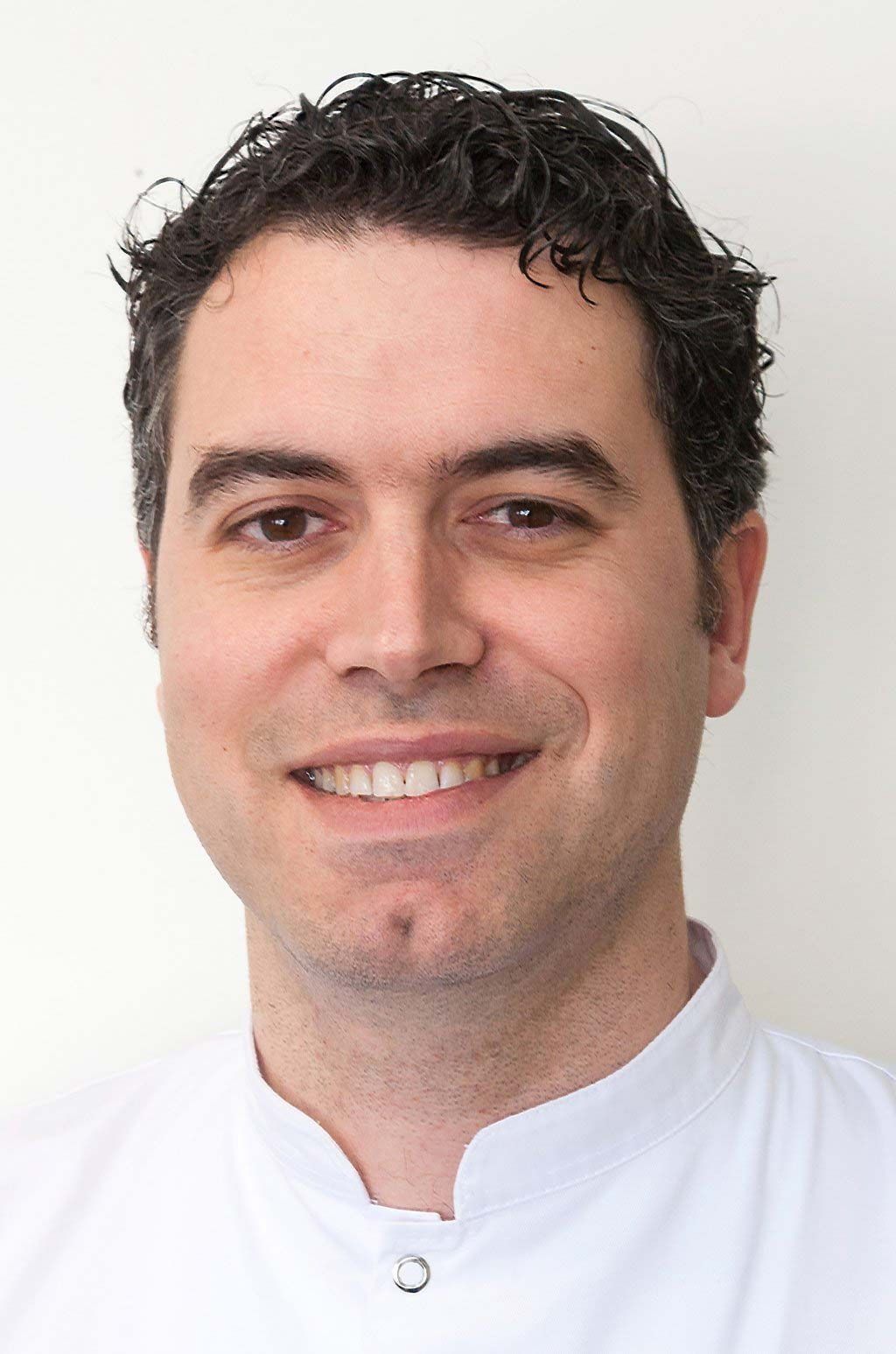 Una persona con cabello oscuro y rizado sonriendo, vistiendo una camisa blanca con cuello sobre un fondo blanco liso.