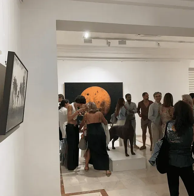 Personas en una galería de arte, reunidas frente a una pintura del sol; una escultura en el medio.