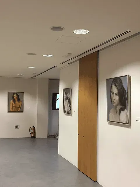 Interior de galería con retratos en paredes blancas, puerta de madera, extintor y suelo gris.