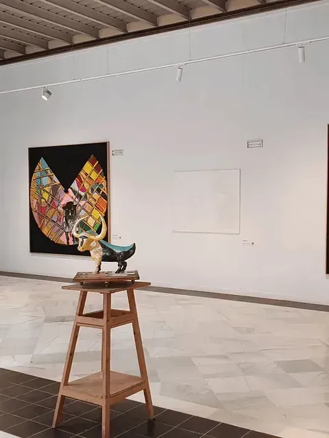 Galería de arte con una escultura sobre soporte de madera, pinturas sobre paredes blancas y suelo de mármol.