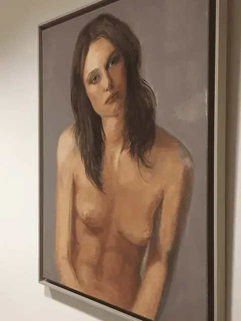 Retrato desnudo de una mujer con cabello castaño y expresión neutra, sobre un fondo gris.