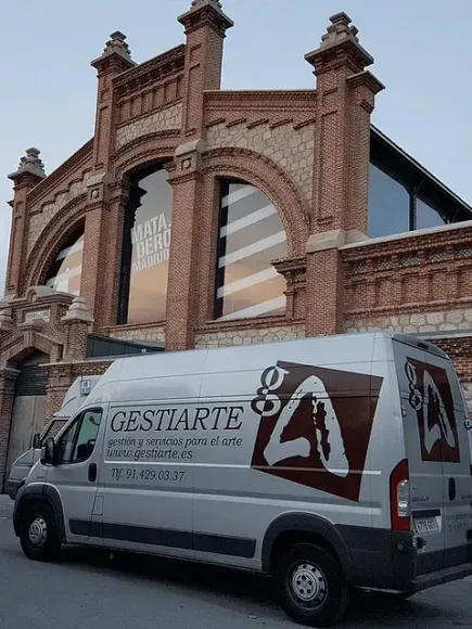 Furgoneta plateada con el logotipo de Gestiarte estacionada afuera de un edificio de ladrillo con ventanas arqueadas.