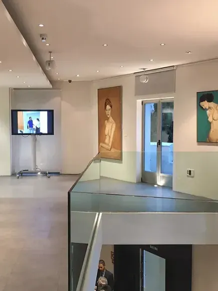 Interior de una galería de arte con pinturas en las paredes, una pantalla de televisión sobre un soporte, una barandilla de vidrio y una persona bajando las escaleras.