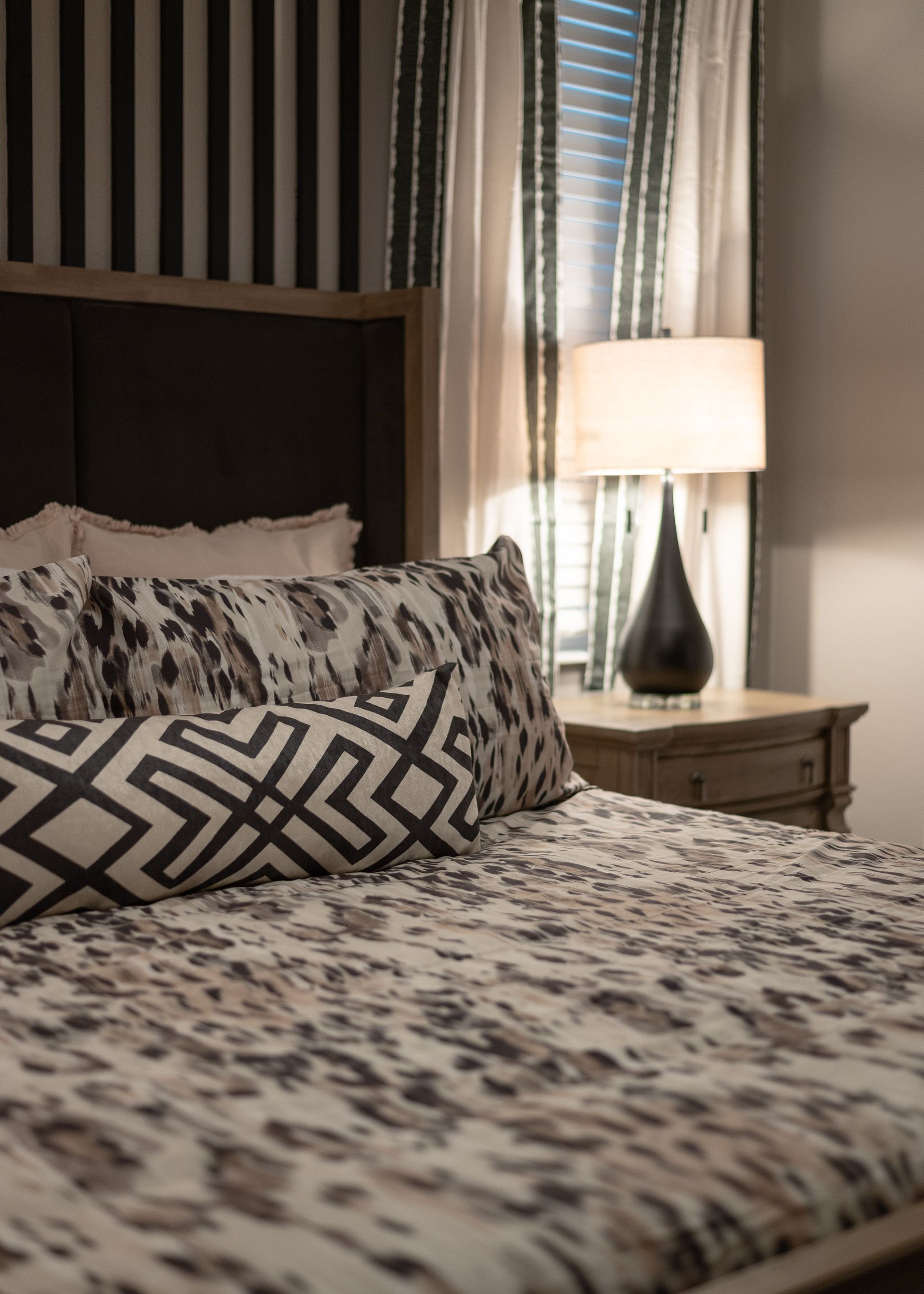 Bedroom with patterned bedding, a lamp on a nightstand, and striped wall behind the bed.