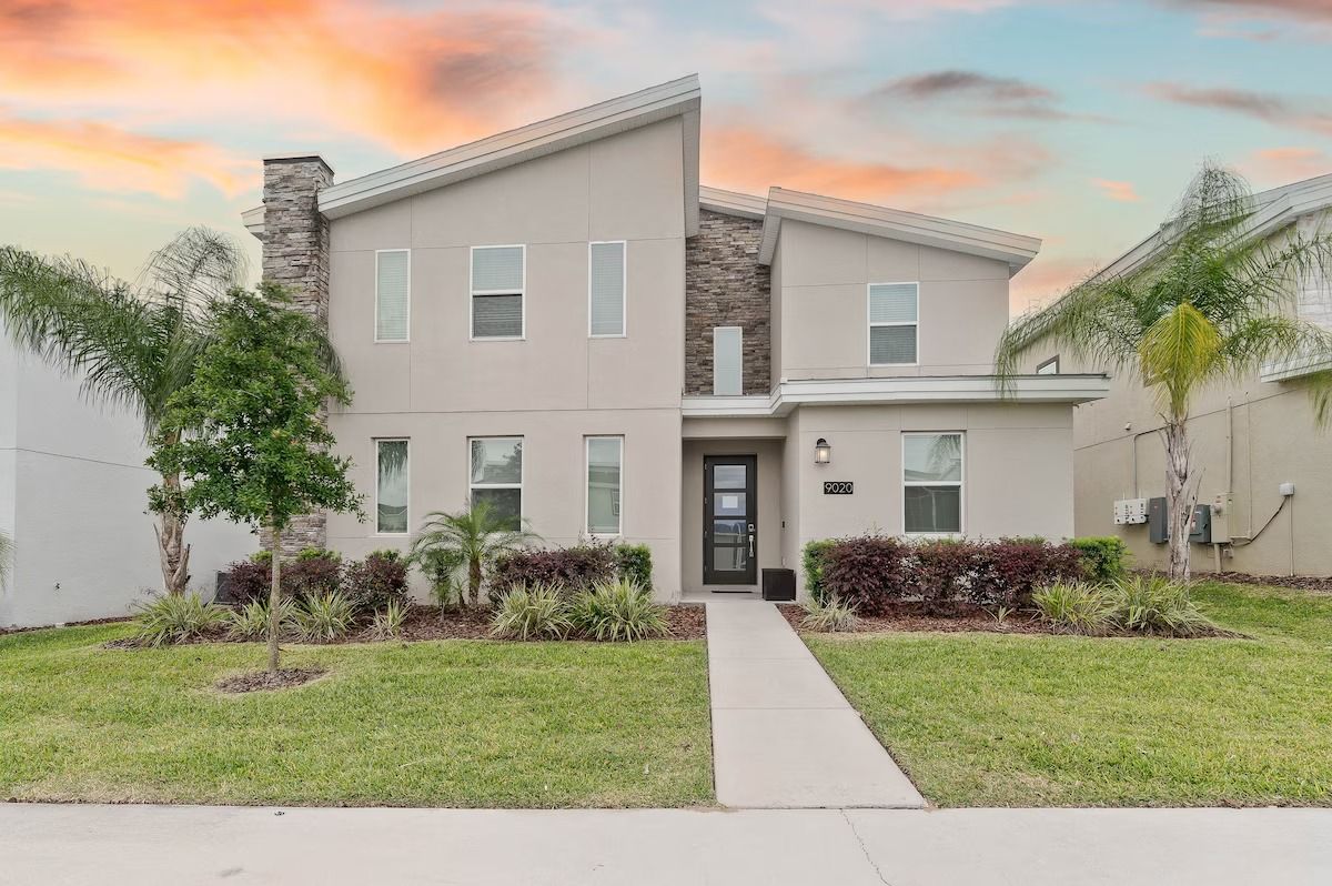 Two-story beige house with a stone accent, sidewalk, and green lawn, under a colorful sunset.