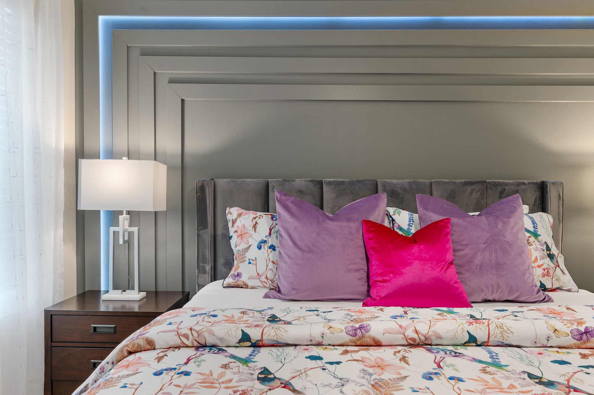 Bedroom with gray headboard, lamp on nightstand, bed with floral duvet and purple pillows.