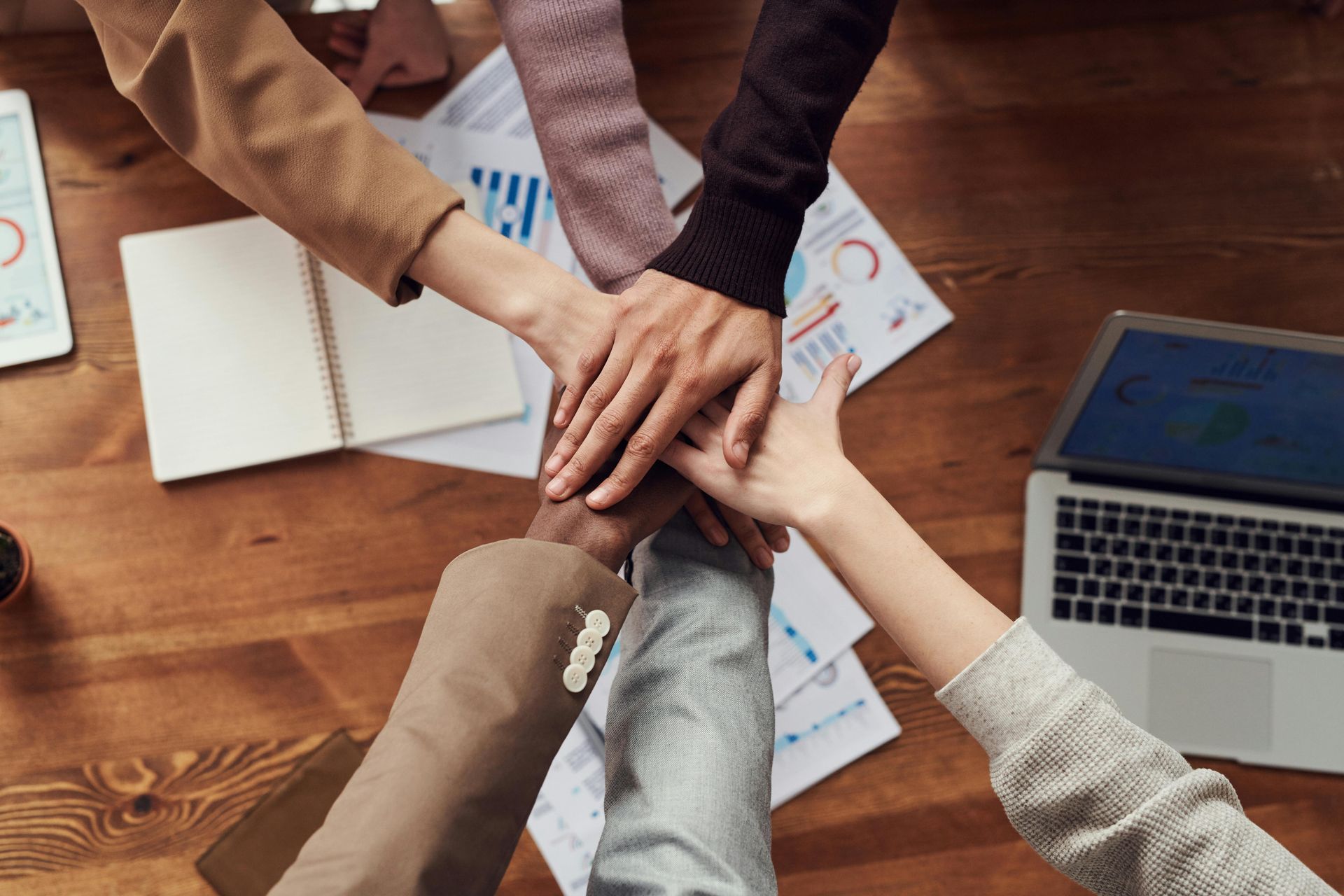 Hands stacked in a team huddle over a wooden table with paperwork and a laptop.