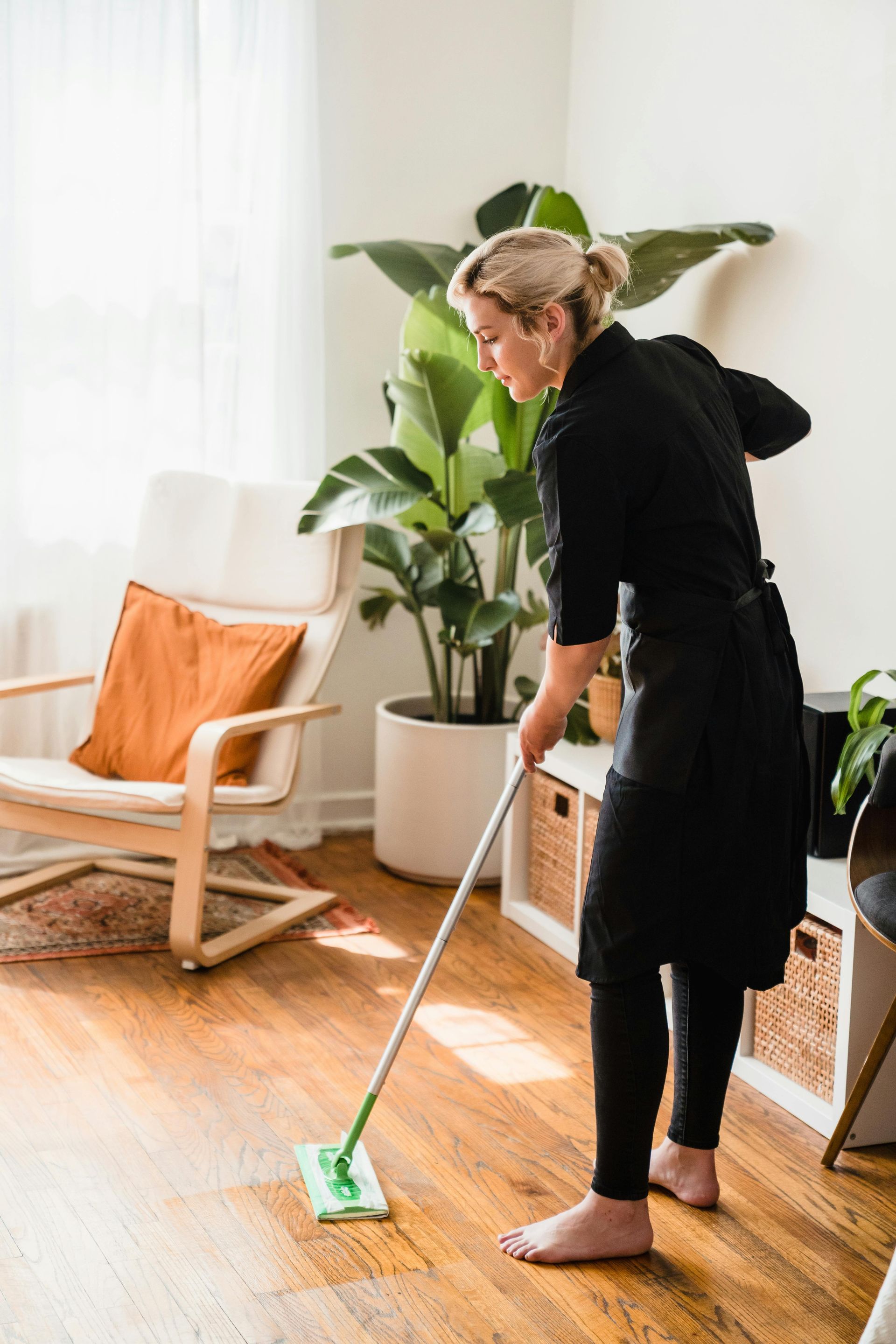 Woman mopping wooden floor in sunlit room; chair, plants, and storage unit visible.