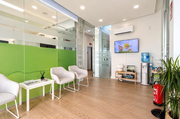 Sala de espera con sillas blancas, mampara verde, suelo de madera, televisor, dispensador de agua y extintor.