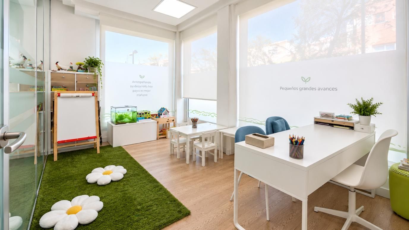 Sala de juegos luminosa con alfombra verde, mesas y sillas blancas, estanterías y grandes ventanales con plantas y flores.