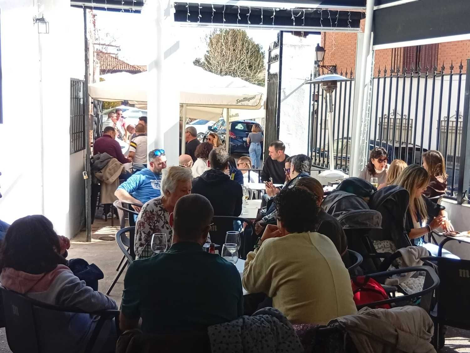 Gente sentada en mesas al aire libre, cenando y socializando bajo un toldo parcial. Día soleado y luminoso.