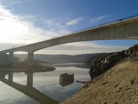 Puente sobre el agua, con estructura de hormigón y una ruina sumergida. Cielo azul y reflejos.