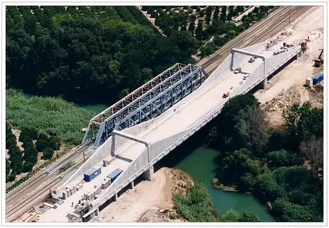 Construcción de un puente sobre un río y una vía férrea. Estructura de hormigón y acero, con contenedores azules visibles.