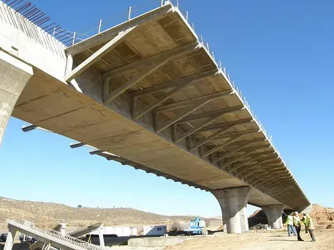 Puente en construcción con soportes de hormigón, cerchas y tablero contra un cielo azul.