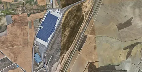 Vista aérea de un gran edificio industrial con techo azul oscuro, situado en un entorno rural y agrícola.