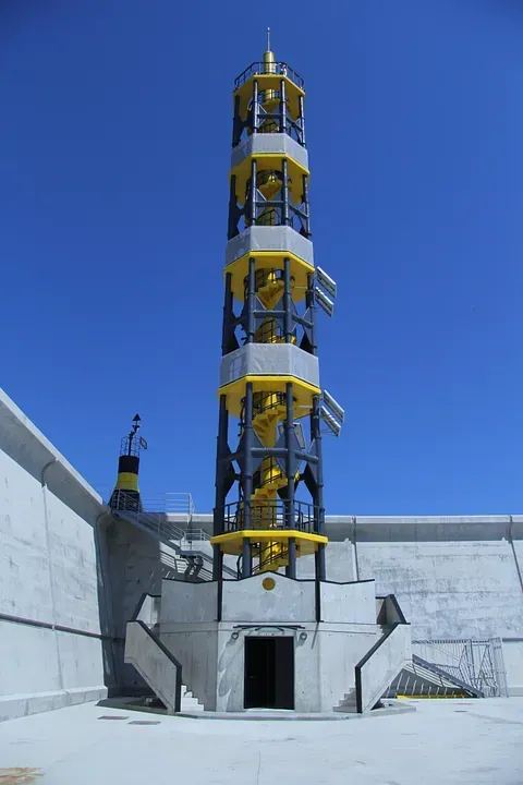 Un faro alto, amarillo y gris contra un cielo azul, construido sobre una estructura de hormigón.