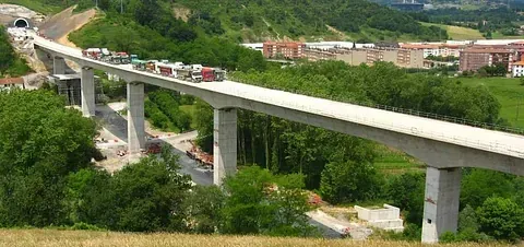 Un largo puente de hormigón con múltiples camiones cruzando sobre frondosos árboles verdes y edificios en la distancia.