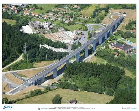 Vista aérea de una larga carretera elevada con pilares altos, que conduce a la entrada de un túnel en una zona boscosa.