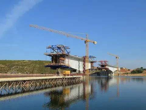 Puente en construcción sobre el agua, grúas, cielo azul y andamios.
