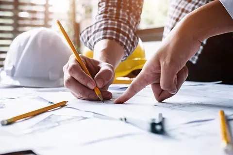 Arquitectos señalando y dibujando sobre planos, rodeados de cascos y lápices.