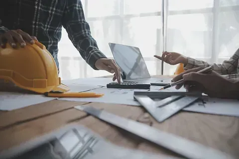 Arquitectos en un escritorio, usando una calculadora, una computadora portátil y planos, discutiendo un proyecto de construcción.