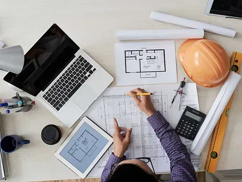 Vista aérea de una persona dibujando planos arquitectónicos en un escritorio con una computadora portátil, una tableta, planos y un casco.