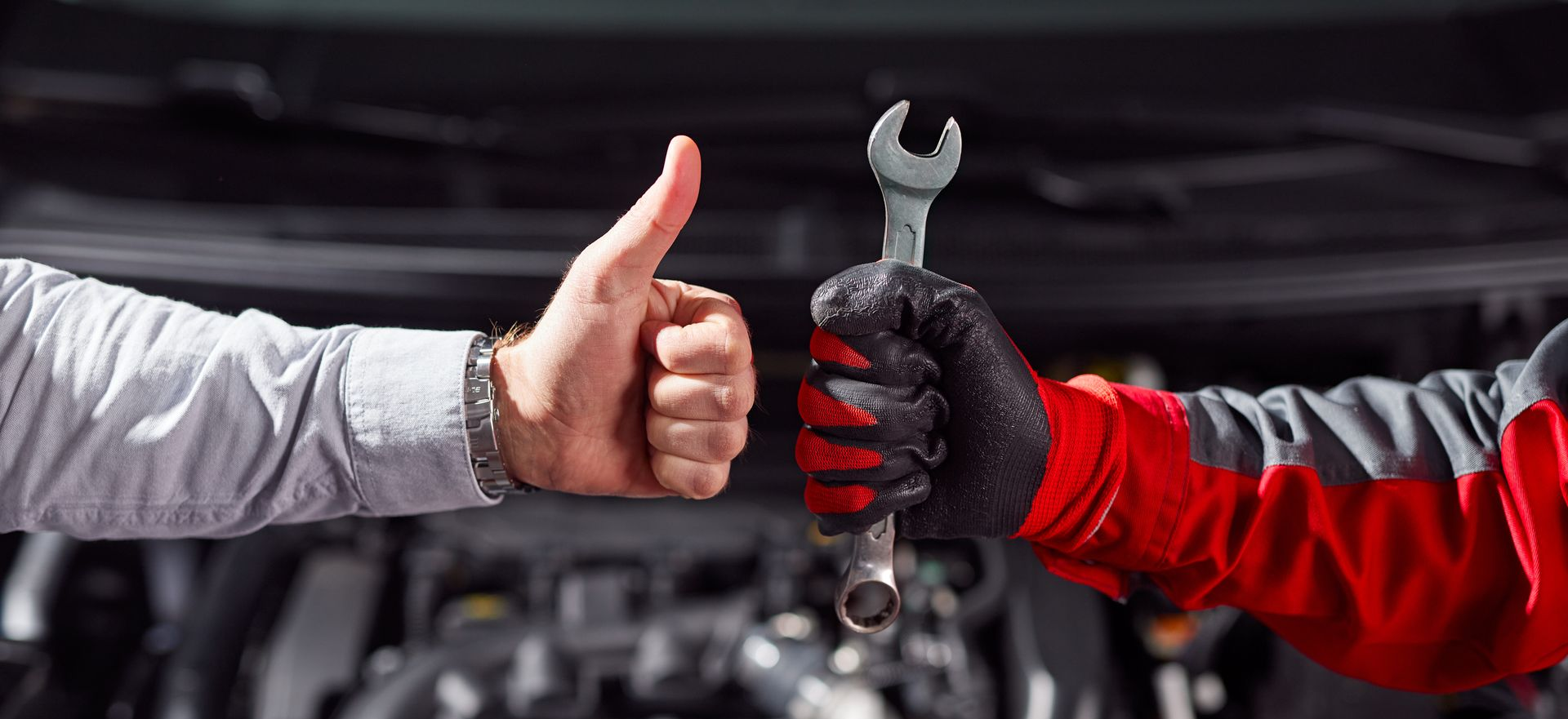 Pouce levé, mécanicien tenant une clé, moteur de voiture en arrière-plan.