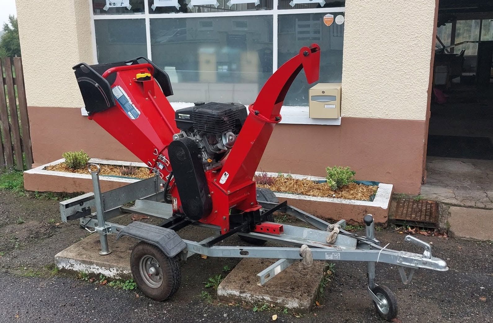 Broyeur de branches rouges sur une remorque à roues, stationné devant un bâtiment.