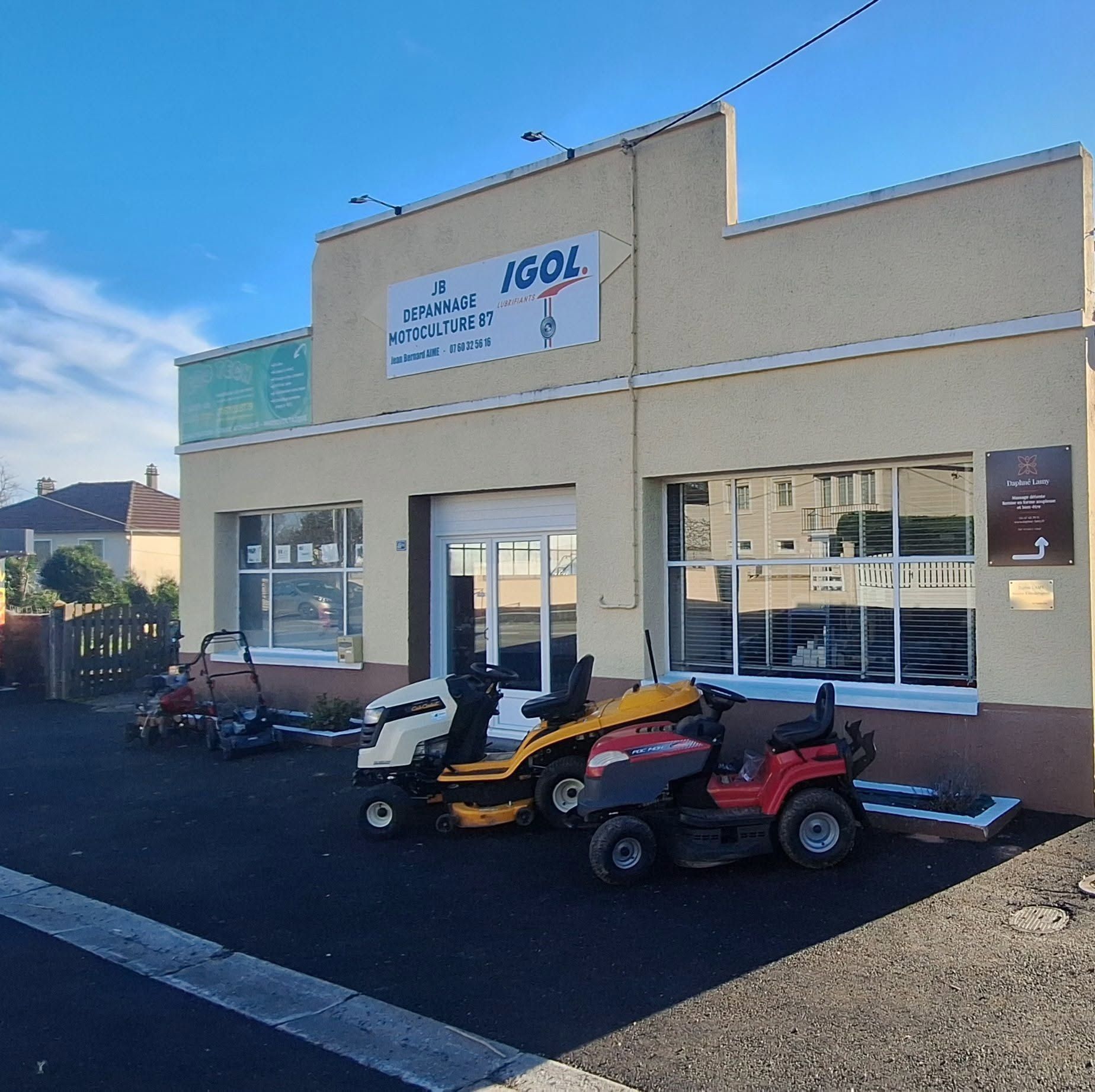 Petite boutique avec une enseigne, du matériel motorisé d'extérieur et une tondeuse autoportée rouge garée devant une rue ensoleillée.